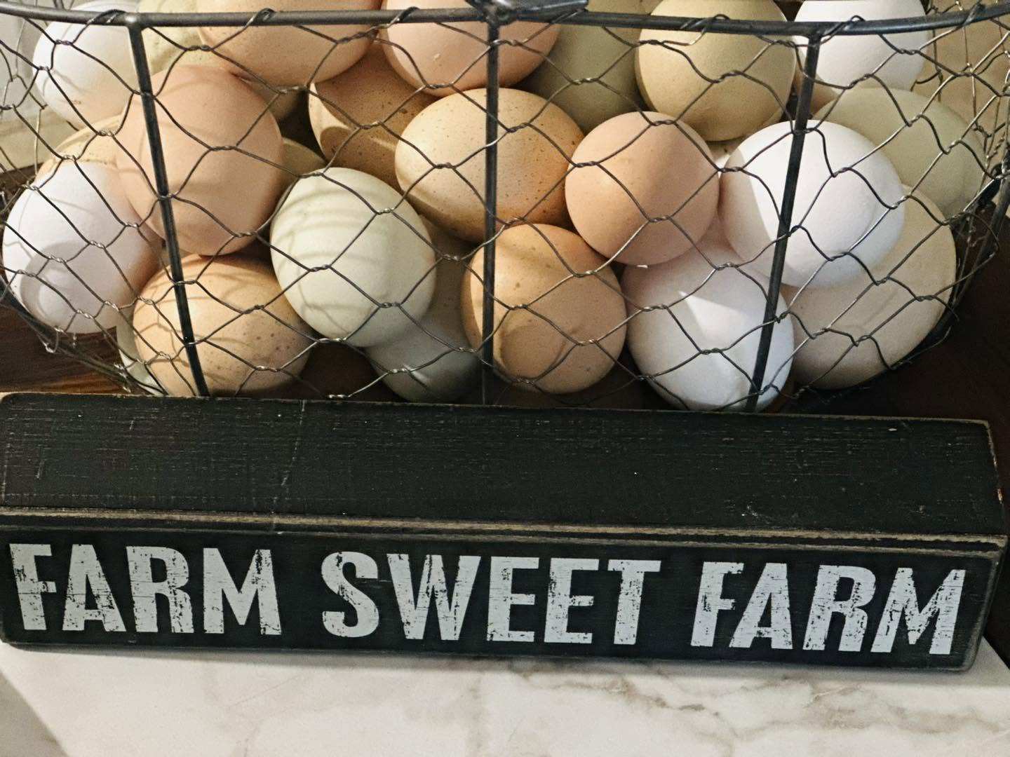 A wire basket of brown, white and pale green farm-fresh eggs on a black "FARM SWEET FARM" sign.
