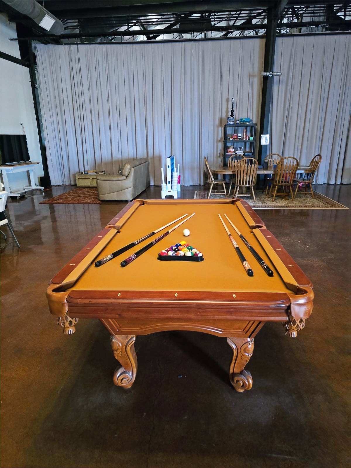 The game room in the community center: a pool table with gold felt, cues and racked balls, a sitting area and dining table behind.