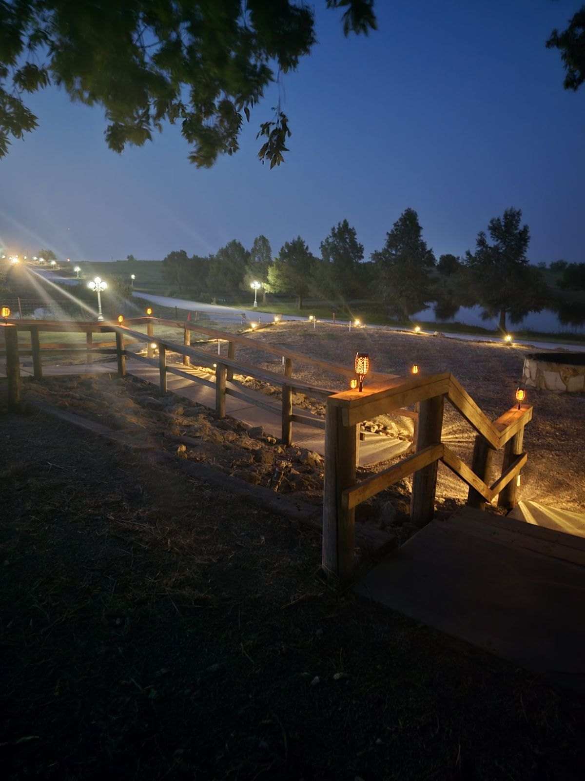 Wooden rails and steps by the lake lit by small lanterns at dusk, with lamp posts along the drive beyond.