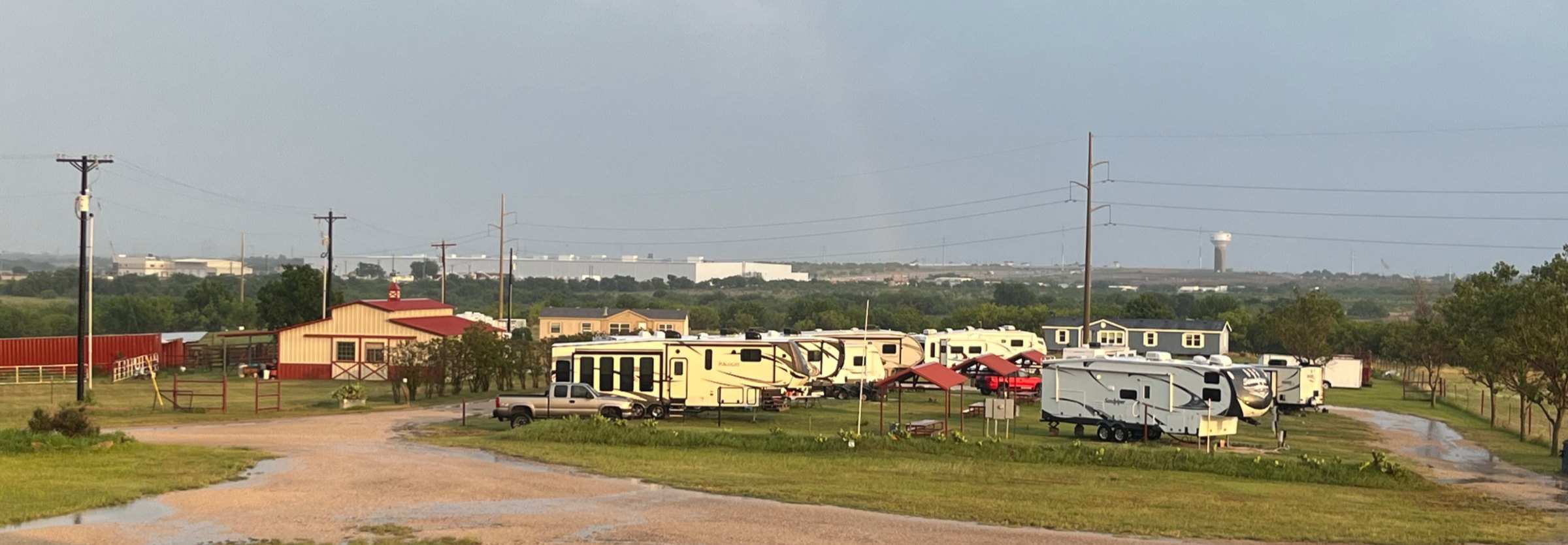 Fifth-wheels and travel trailers parked along the grass in the RV area, with covered picnic tables between the pads and the Denton water tower on the horizon.