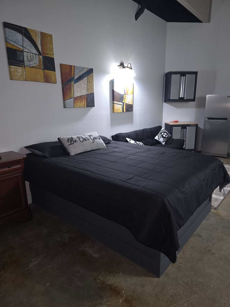 Studio 201: a bed with a black comforter and a black futon under three abstract paintings, a fridge in the far corner.