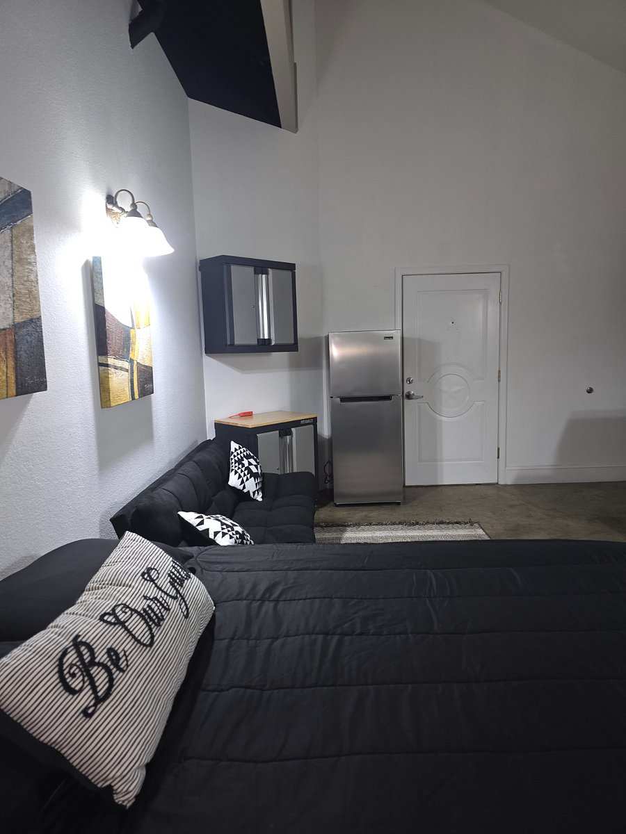 Studio 201 from the bed: a “Be Our Guest” pillow on a black comforter, the futon, the fridge and the entry door.