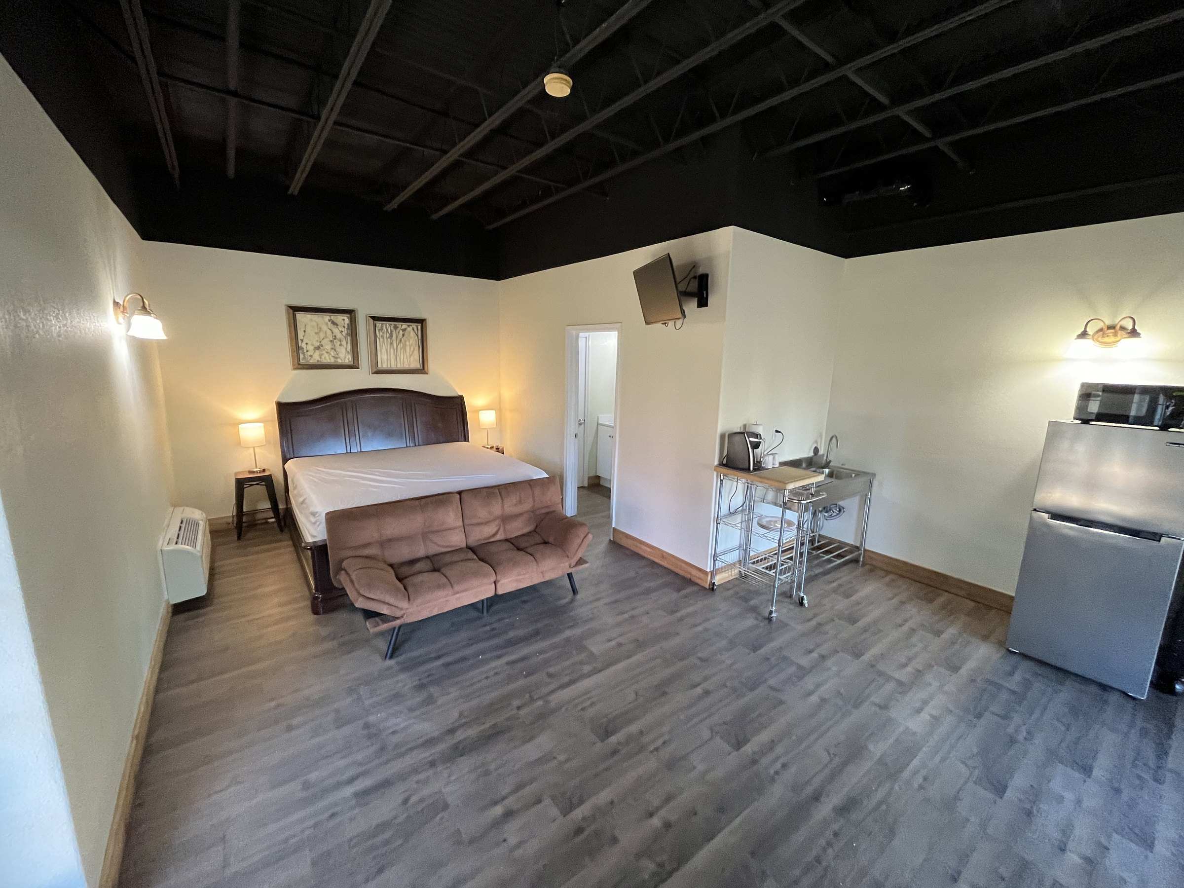A studio with wood-look floors under a black ceiling: a bed and loveseat, a wall TV, a kitchenette cart and a stainless fridge.